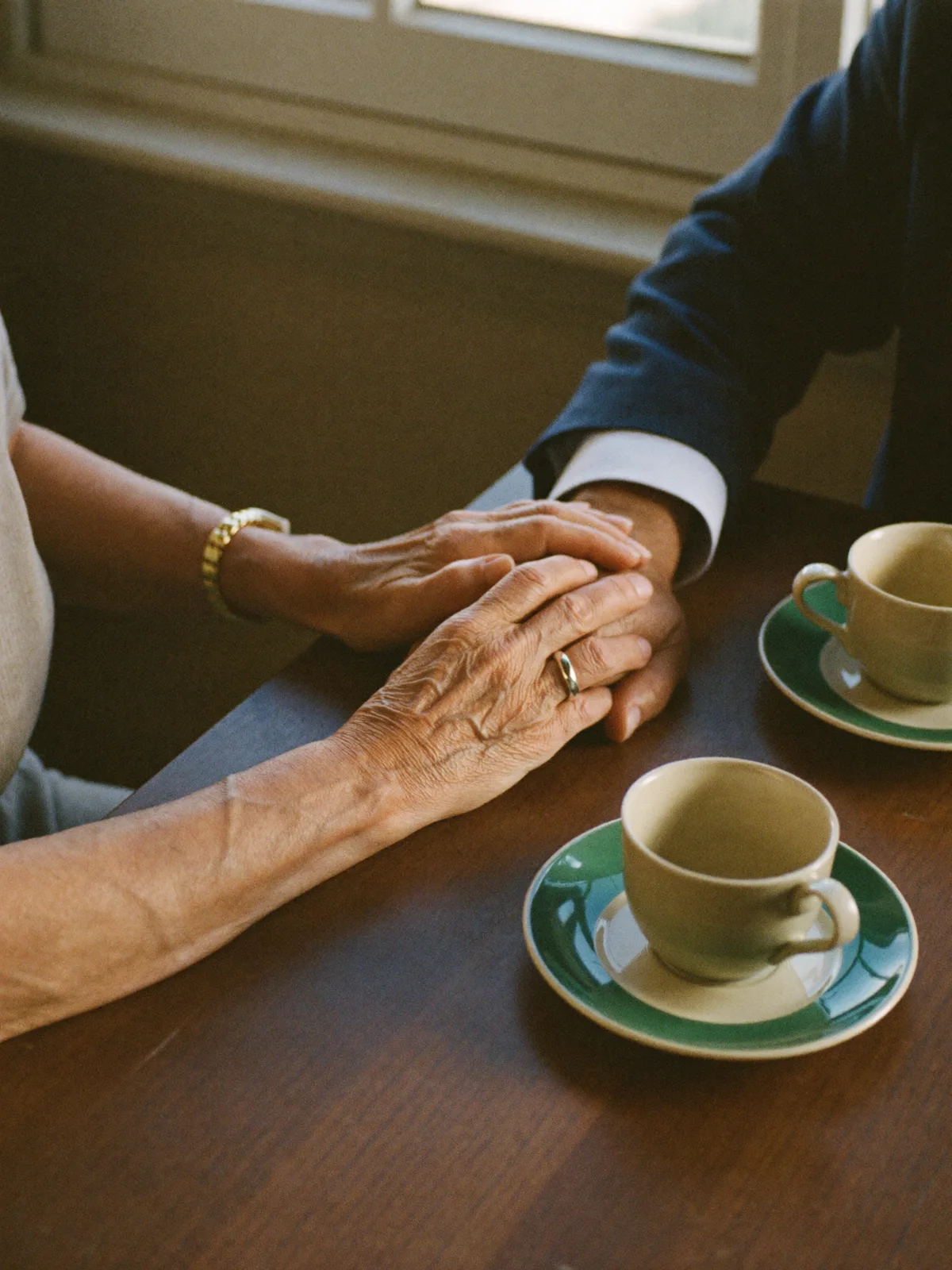 An older couple's hands resting together at a kitchen table in morning light